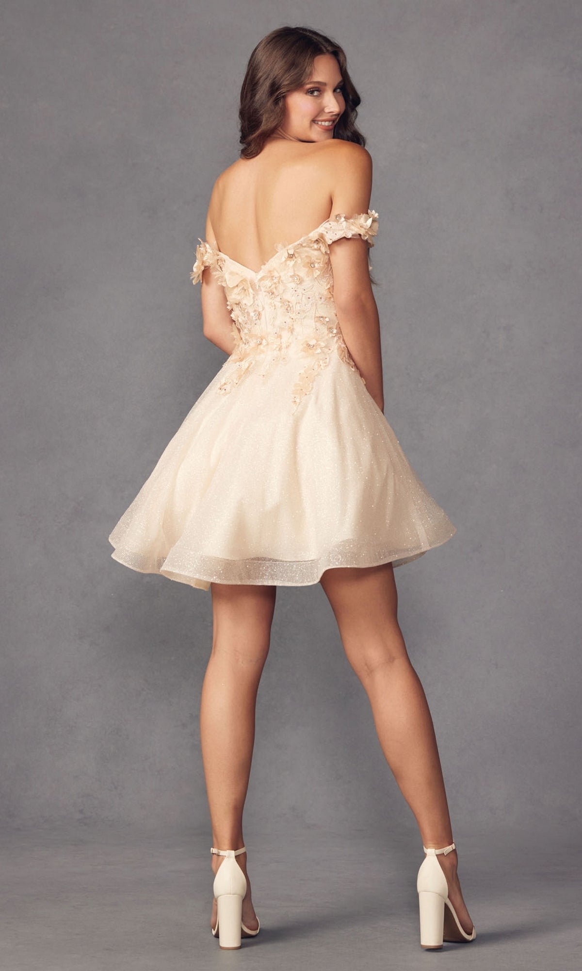 A woman in the Floral Off-the-Shoulder Short Homecoming Dress 861 and white heels stands with her back turned, glancing over her shoulder.